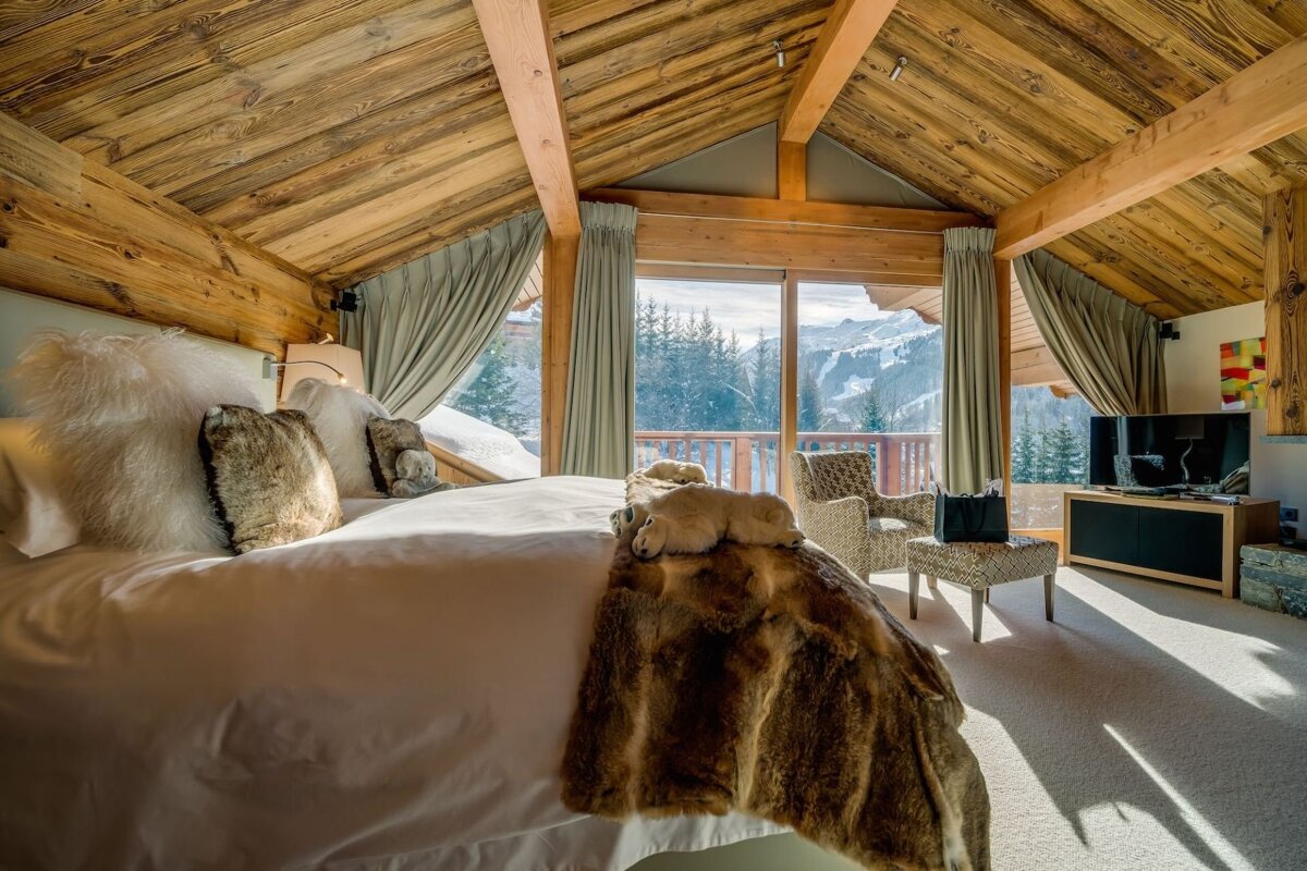 Cozy ski chalet bedroom with vaulted wooden ceilings, a plush bed, and large windows revealing panoramic views of snowy mountains.