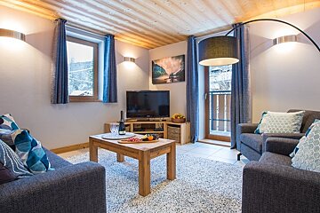 A cozy chalet living room featuring grey sofas, a wooden coffee table, and windows showing a snowy winter landscape.