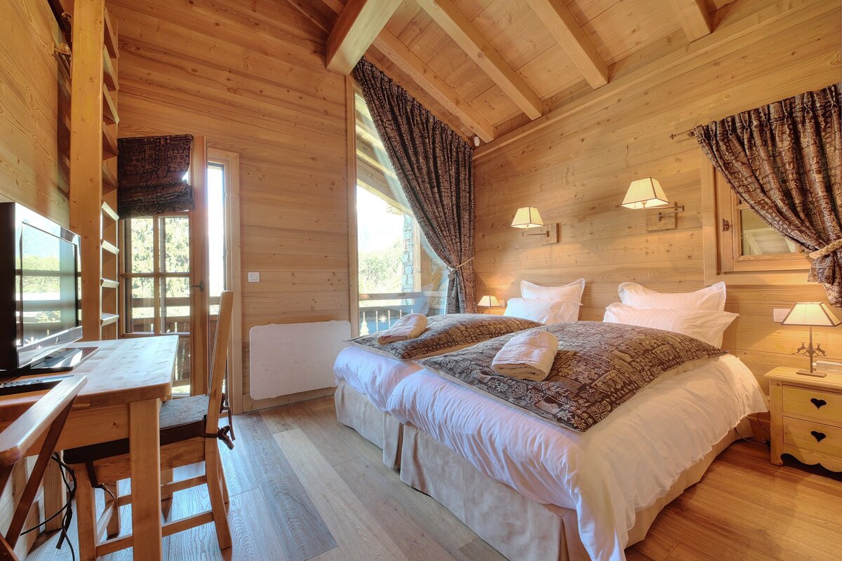 A cozy chalet bedroom with light wooden walls/ceiling, two beds, a desk with TV, and large windows with patterned curtains.