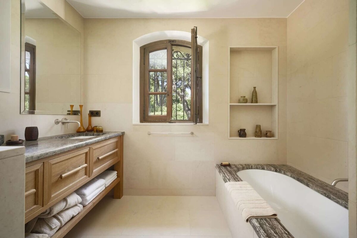 An elegant, light-toned bathroom with a wooden vanity, marble-rimmed bathtub, and an arched window offering a view of lush trees.
