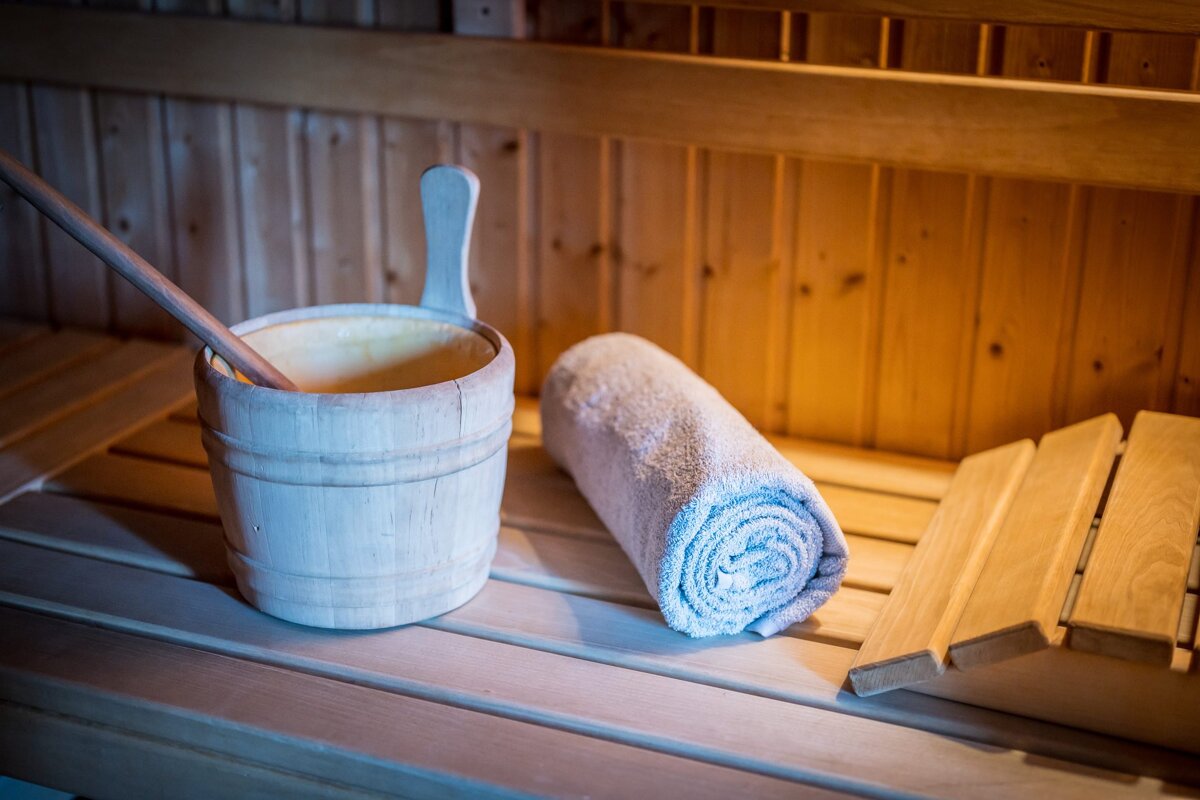 A wooden sauna bucket with a ladle, a rolled-up towel, and a wooden headrest sit on a bench inside a cozy sauna.