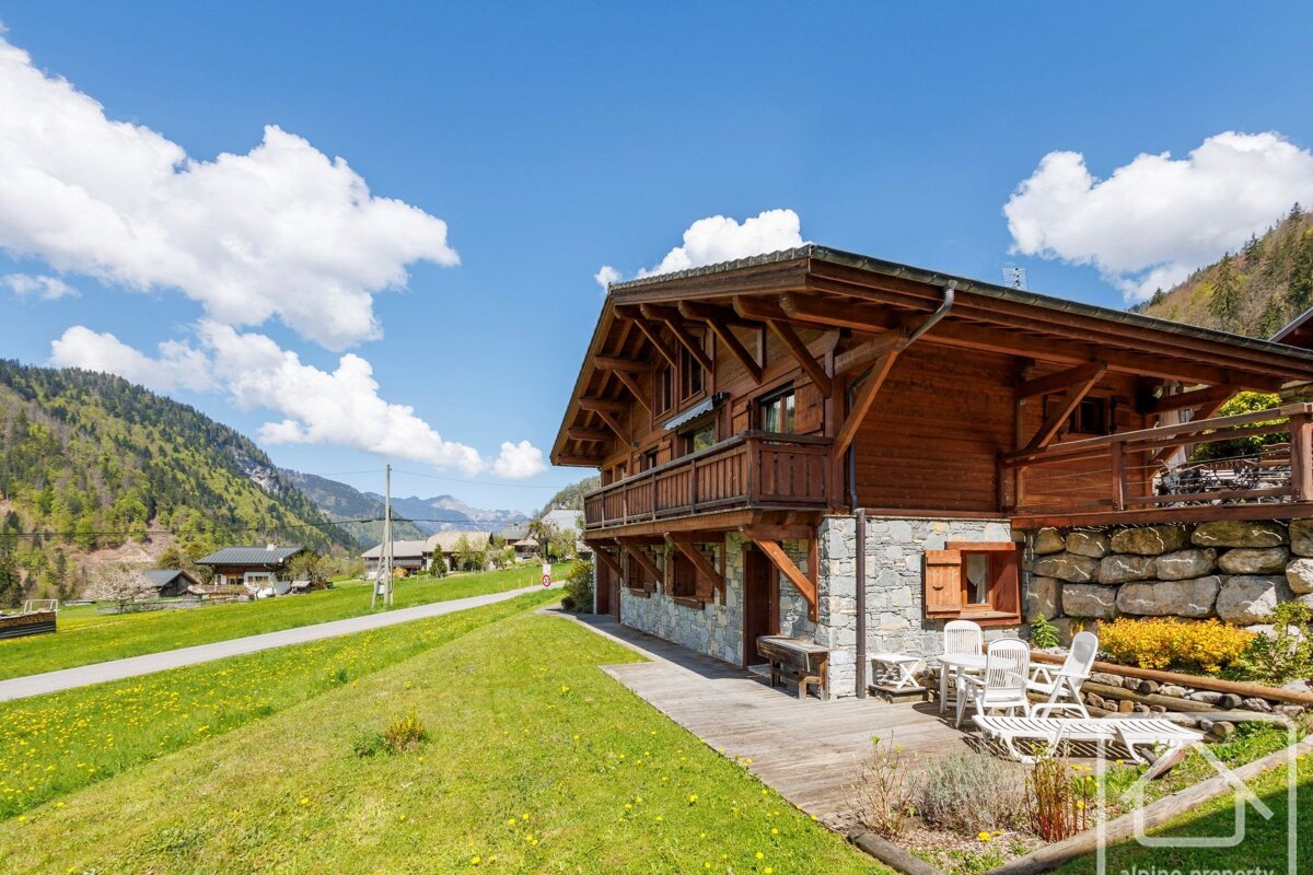 A charming wooden alpine chalet with a patio sits in a lush green valley, surrounded by mountains under a vibrant blue sky with fluffy white clouds.