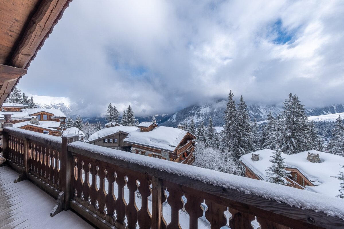 A snowy balcony overlooks an alpine village with traditional chalets, snow-dusted pine trees, and cloudy mountain peaks under a winter sky.