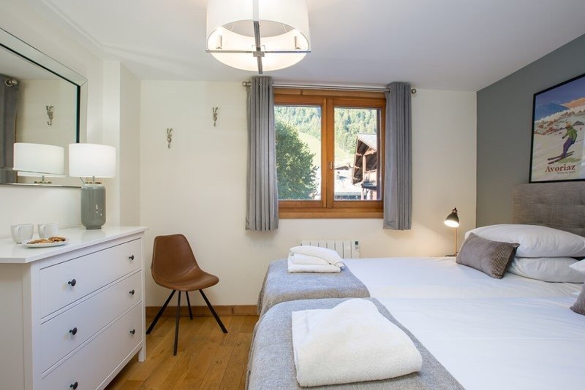 Cozy chalet bedroom featuring twin beds, a white dresser, a brown chair, and a window looking out at mountain scenery.