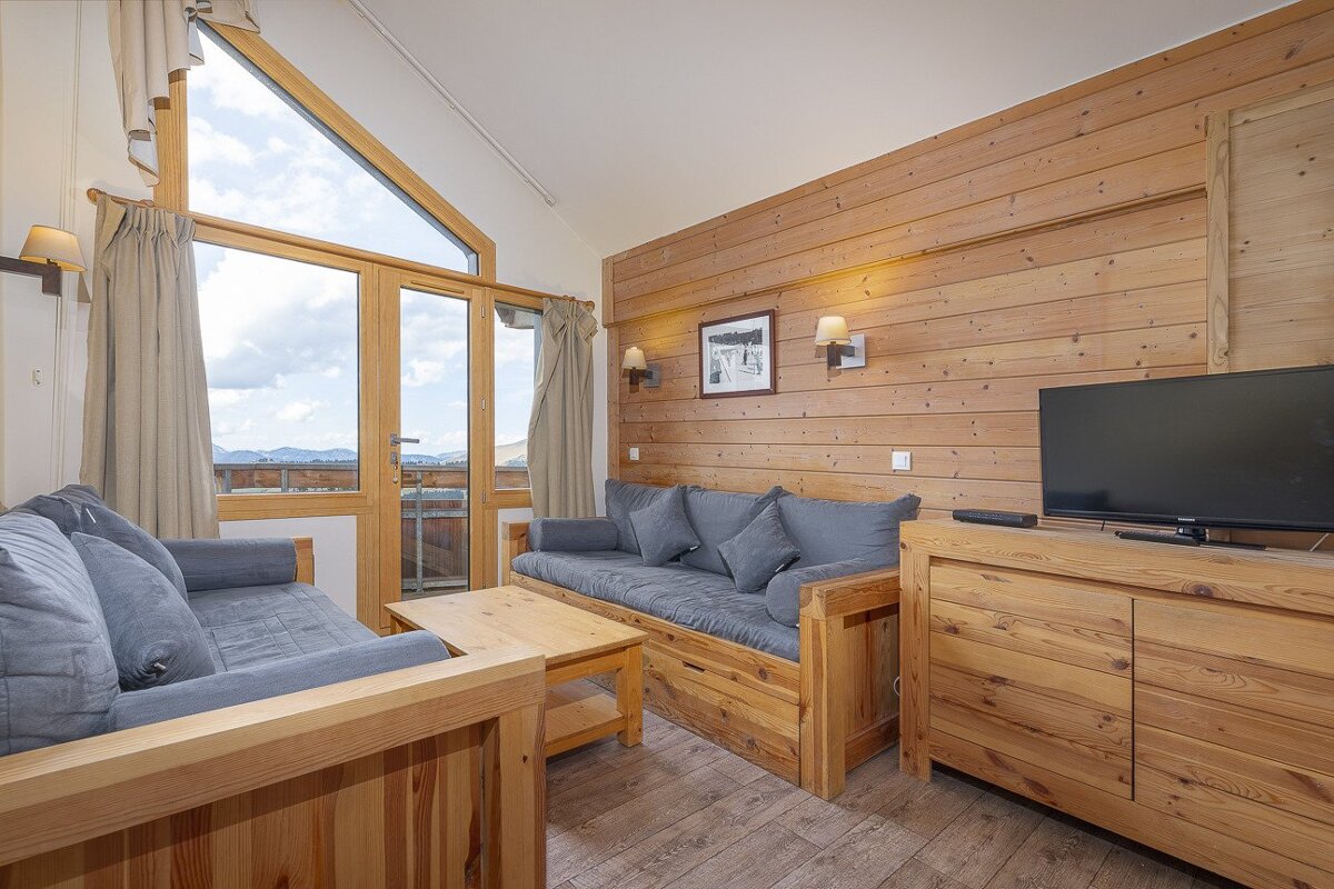 A cozy, wood-paneled living room with blue sofas, a TV, and a large window offering a beautiful mountain view and access to a balcony.
