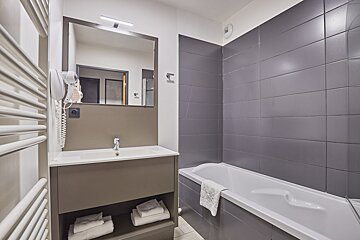 Modern bathroom featuring a sink vanity with mirror and hairdryer, a towel warmer, and a bathtub with sleek gray tiled walls.