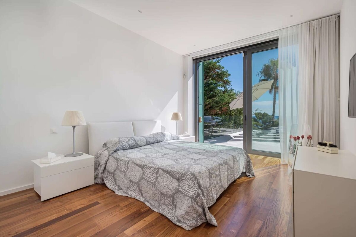 A bright, modern bedroom features a bed, wooden floor, and white walls. A large glass door overlooks a sunny patio with trees and a glimpse of the sea.