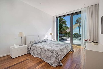 A bright, modern bedroom features a bed, wooden floor, and white walls. A large glass door overlooks a sunny patio with trees and a glimpse of the sea.