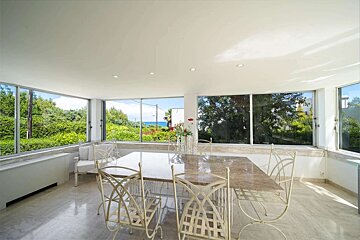 Bright room with panoramic windows showing lush greenery and a glimpse of the ocean. A dining table and white chairs are on a marble floor.
