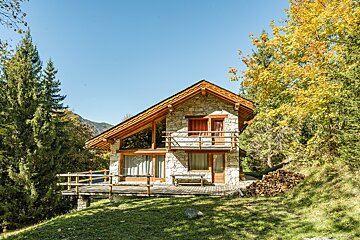 A rustic stone and wood chalet with a deck on a grassy hill, surrounded by trees, with mountains in the distance under a clear sky.