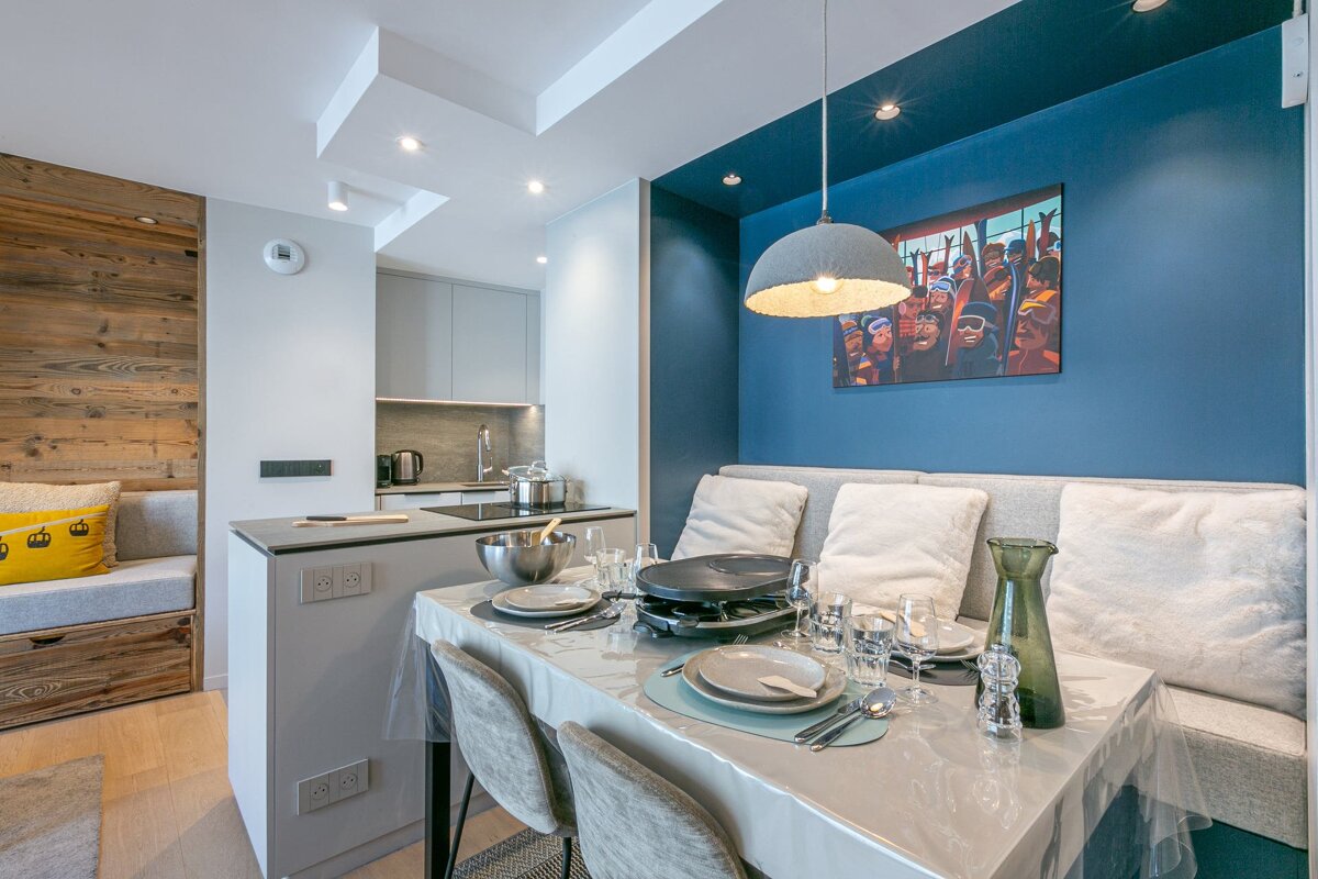 A modern kitchen-dining area with a blue accent wall, art, and a raclette grill on the table, plus wood-paneled seating nearby.