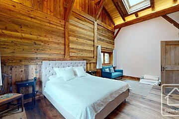 A rustic-modern bedroom featuring warm wooden walls, floor, and vaulted ceiling with a skylight. A large bed and a blue sofa complete the cozy space.