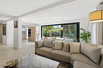A bright, modern open-plan living room with a neutral sofa, glass coffee table, large windows to a lush garden, and a visible dining area/kitchen.