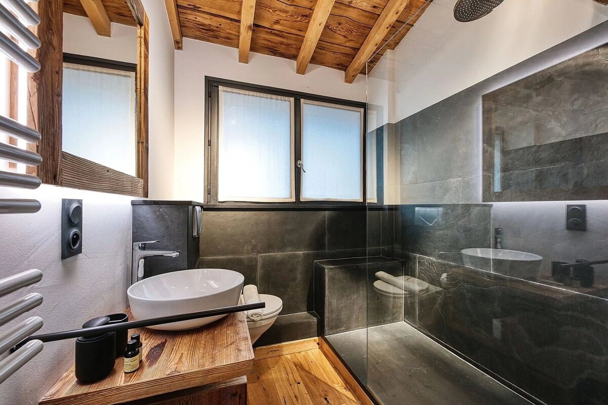 Modern bathroom with a rustic touch, featuring dark tiled walls, exposed wooden ceiling beams, a wooden vanity, and a glass shower.