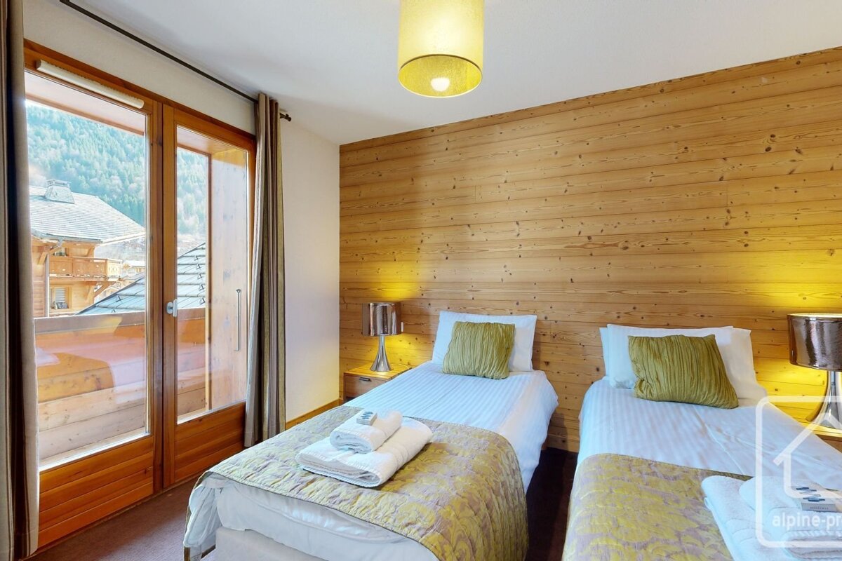 Cozy twin bedroom with a wooden panel accent wall, bedside lamps, green pillows, and a balcony offering a view of a mountain village.