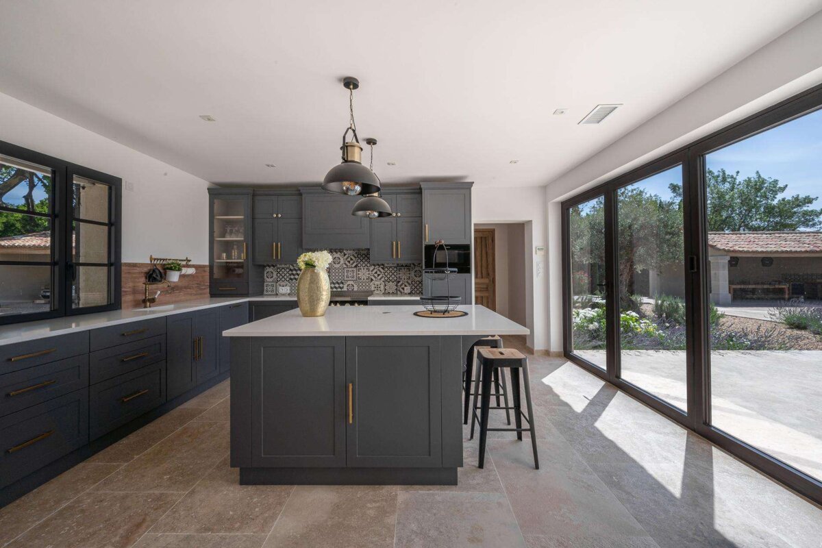 A sleek dark gray kitchen featuring a white island, modern pendant lights, and expansive glass doors opening to a bright outdoor patio.