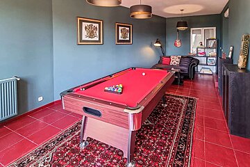 A vibrant game room with a red felt pool table, dark teal walls, and bold red tiled floors. A black sofa, shelves, and a wall-hung guitar complete the scene.