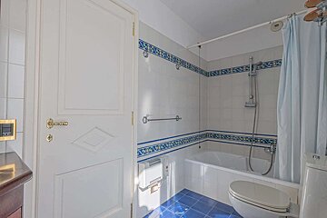 A bathroom features white tiled walls with blue and yellow decorative borders, a blue tiled floor, a bathtub with a shower, and a toilet. A white door with gold hardware is on the left.
