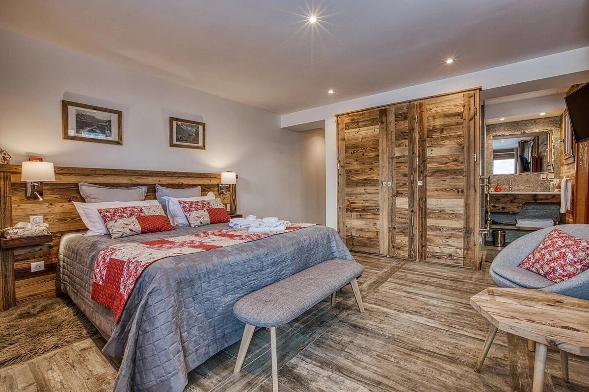 A cozy bedroom with a rustic-modern feel, featuring a large bed with patterned bedding, wooden walls/furniture, and an open ensuite bathroom.