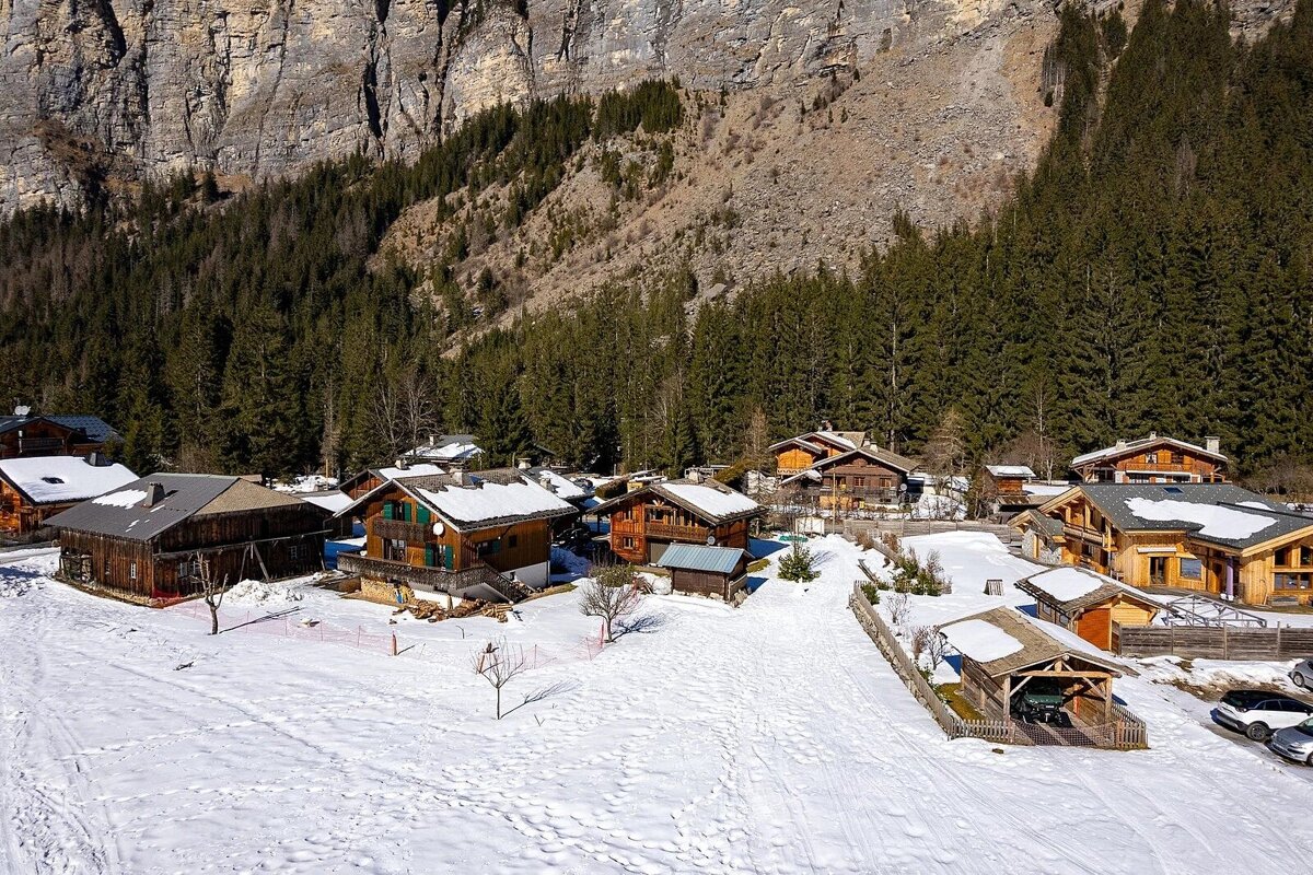 A snowy mountain village with traditional wooden chalets, dense pine forests, and a rocky cliff backdrop under a clear sky.