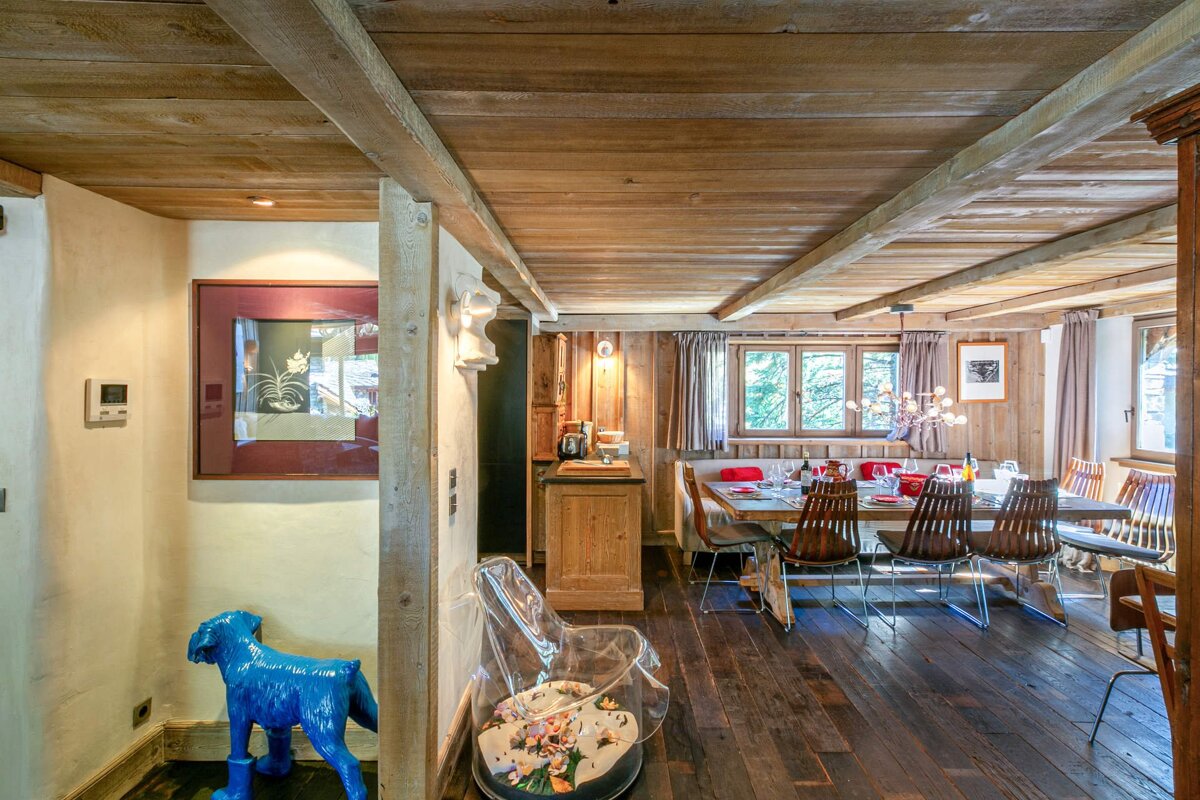 A cozy wooden chalet interior features a rustic dining room with a long table, modern chandelier, and a playful blue dog sculpture.