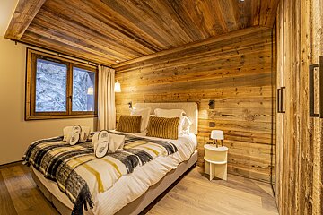 A cozy, rustic bedroom featuring extensive wood paneling on walls and ceiling, a bed with a plaid blanket and slippers, and a window with a view.
