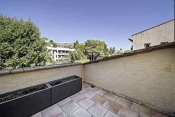 A sunlit balcony with tiled floor and two dark planters, overlooking lush green trees and distant buildings under a clear blue sky.