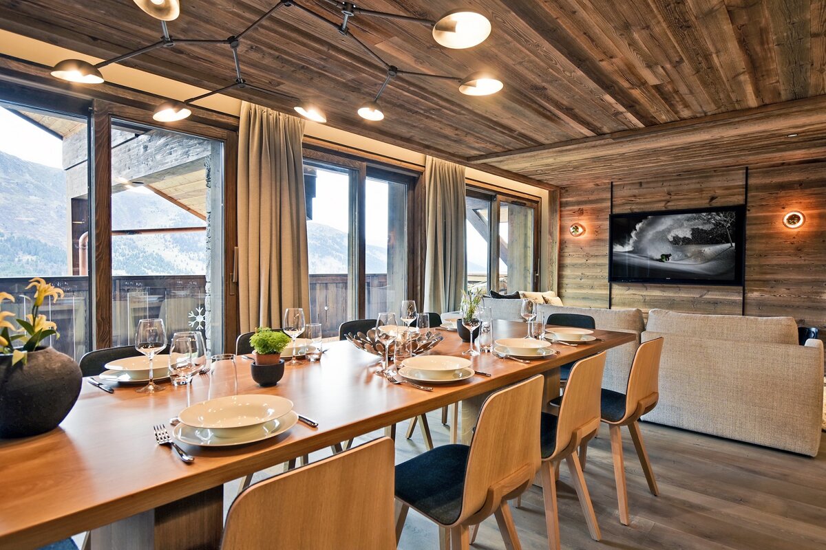 A stylish dining area and living room in a modern chalet, featuring wooden walls/ceiling, a set table, and mountain views from large windows.