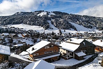 A snowy village nestled in a valley, surrounded by pine-covered mountains with visible ski slopes, under a bright blue sky with scattered clouds.