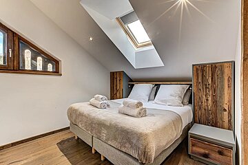 Cozy attic bedroom with a bed, wooden accents, sloped ceilings, and two windows (one triangular, one skylight), creating a warm atmosphere.