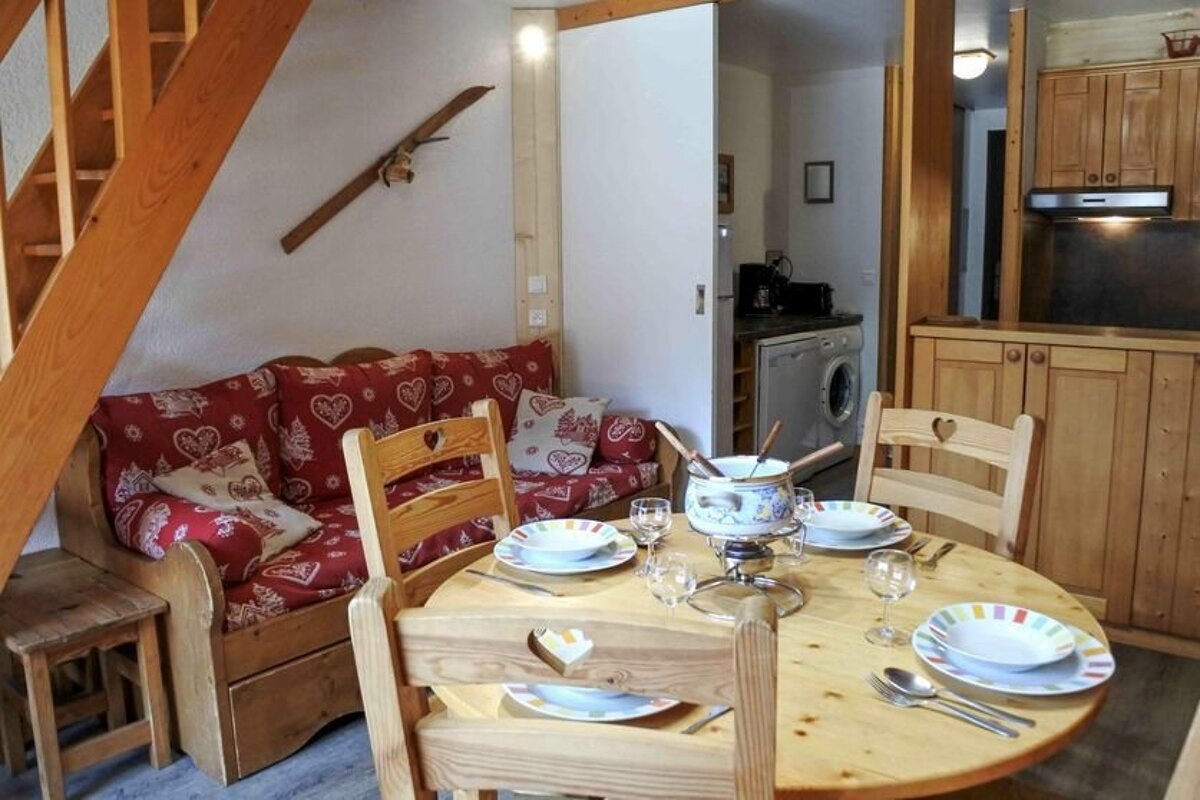 Cozy wooden chalet interior with a fondue pot on a dining table, red heart-patterned sofa, and a compact kitchen. A ski decorates the wall.