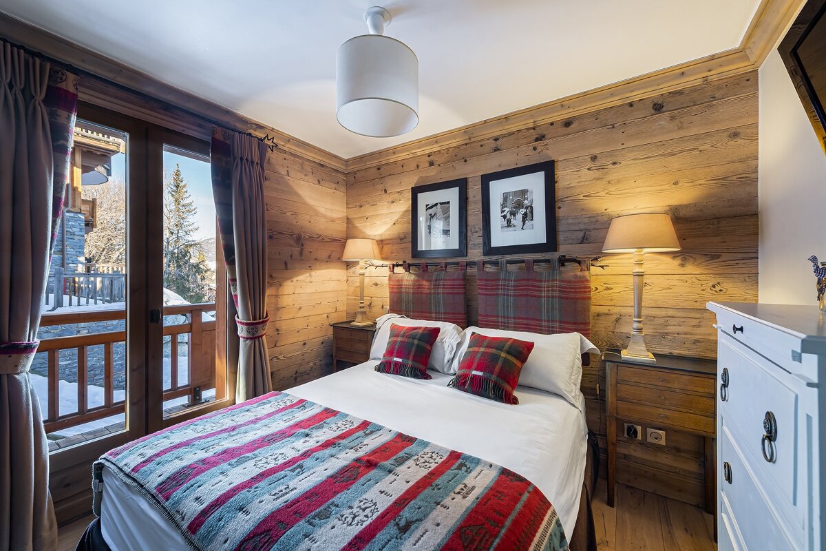 Cozy alpine bedroom with wood-paneled walls, plaid bedding, and a balcony overlooking a snowy mountain landscape.