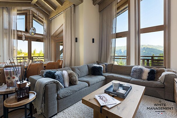 Cozy luxury chalet living room featuring a grey sectional, wooden coffee table, and tall windows showcasing scenic mountain views.