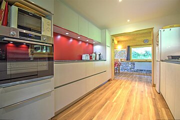 Sleek kitchen with white cabinets, a red accent wall, and wood floor, opening to a rustic dining area with stone walls and wood.
