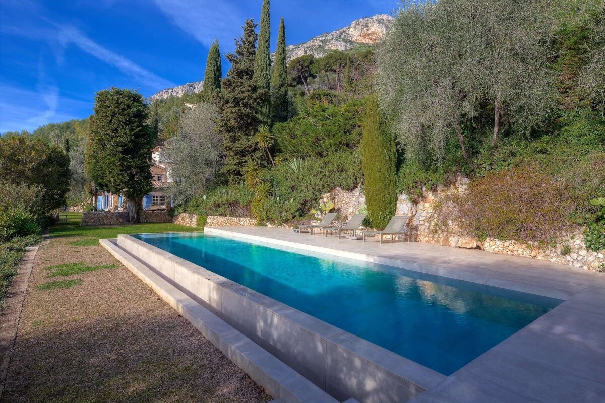 A scenic long swimming pool leads to a charming house nestled among lush trees, with majestic mountains rising under a bright blue sky.