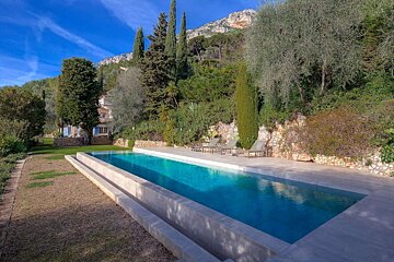 A scenic long swimming pool leads to a charming house nestled among lush trees, with majestic mountains rising under a bright blue sky.