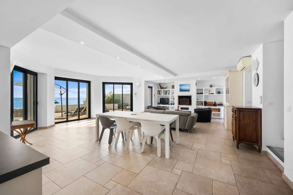 Bright, open-plan living/dining room with large windows offering sea views, light tiled floor, white walls, and built-in shelves.