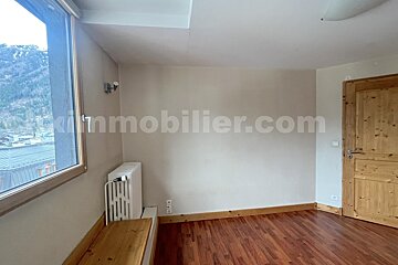 Empty room with wooden floor, light walls, a window overlooking mountains, a radiator, and a wooden door.