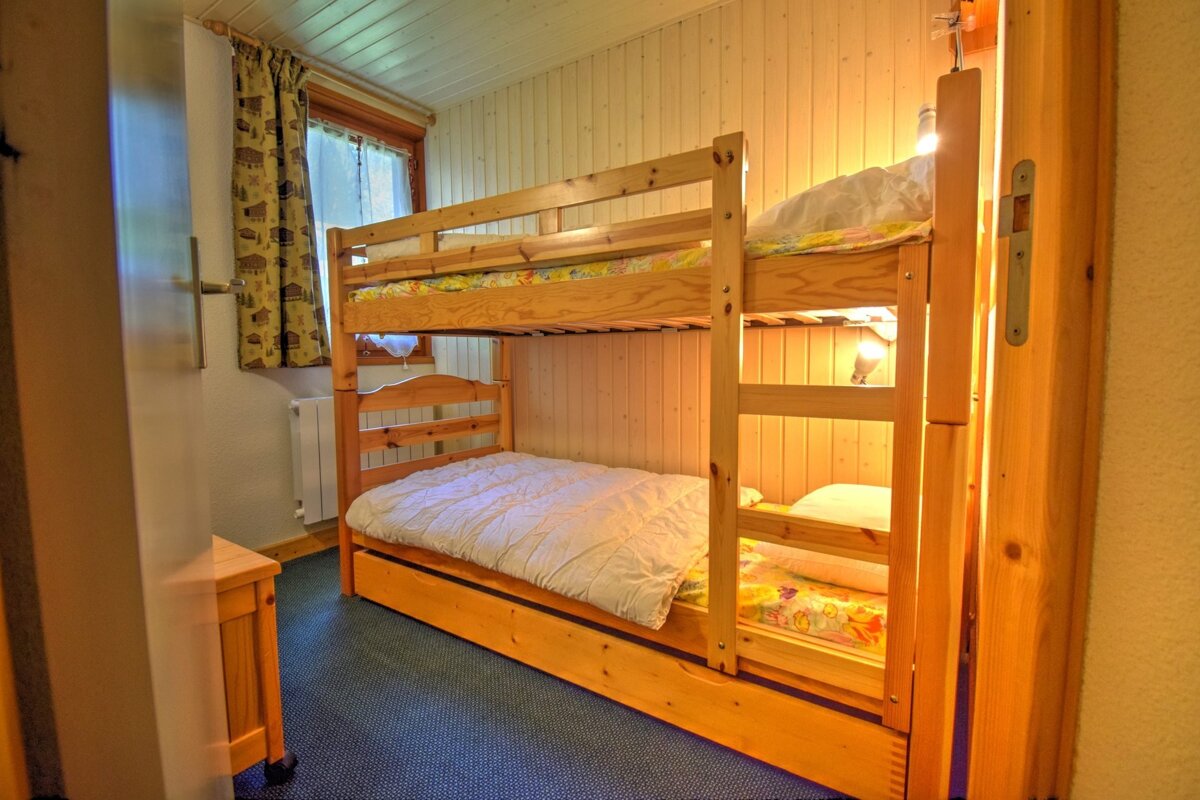 A cozy room with wooden bunk beds, wood-paneled walls, a window with patterned curtains, and blue carpet. Warm lighting illuminates the beds.
