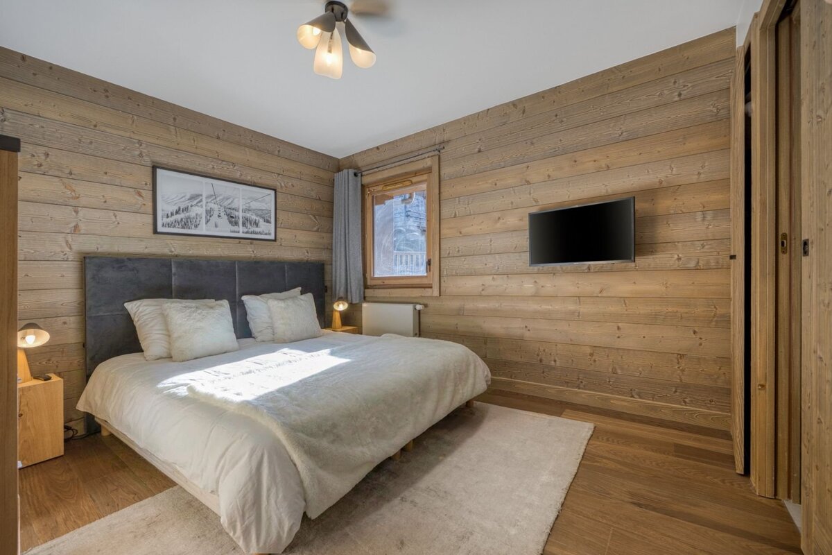 A rustic, modern bedroom featuring wood-paneled walls, a large bed with white linens, a wall-mounted TV, and warm lighting.