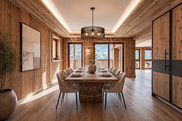 Rustic-modern chalet dining room featuring wood-paneled walls, a long wooden table, elegant chairs, and a stylish dark chandelier.