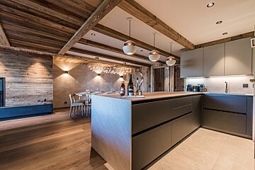 Modern alpine-style kitchen with sleek grey cabinets, wooden beamed ceilings, a large island, and a dining area in the background.