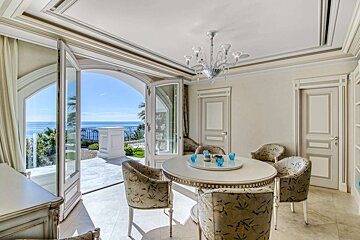 An elegant dining room with a round table, chandelier, and ornate doors, featuring a stunning arched balcony overlooking a sunny, expansive sea.