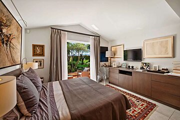 A modern bedroom with a large bed, vaulted ceiling, and open doors to a lush garden and ocean. Features a TV, built-in storage, and earthy decor.