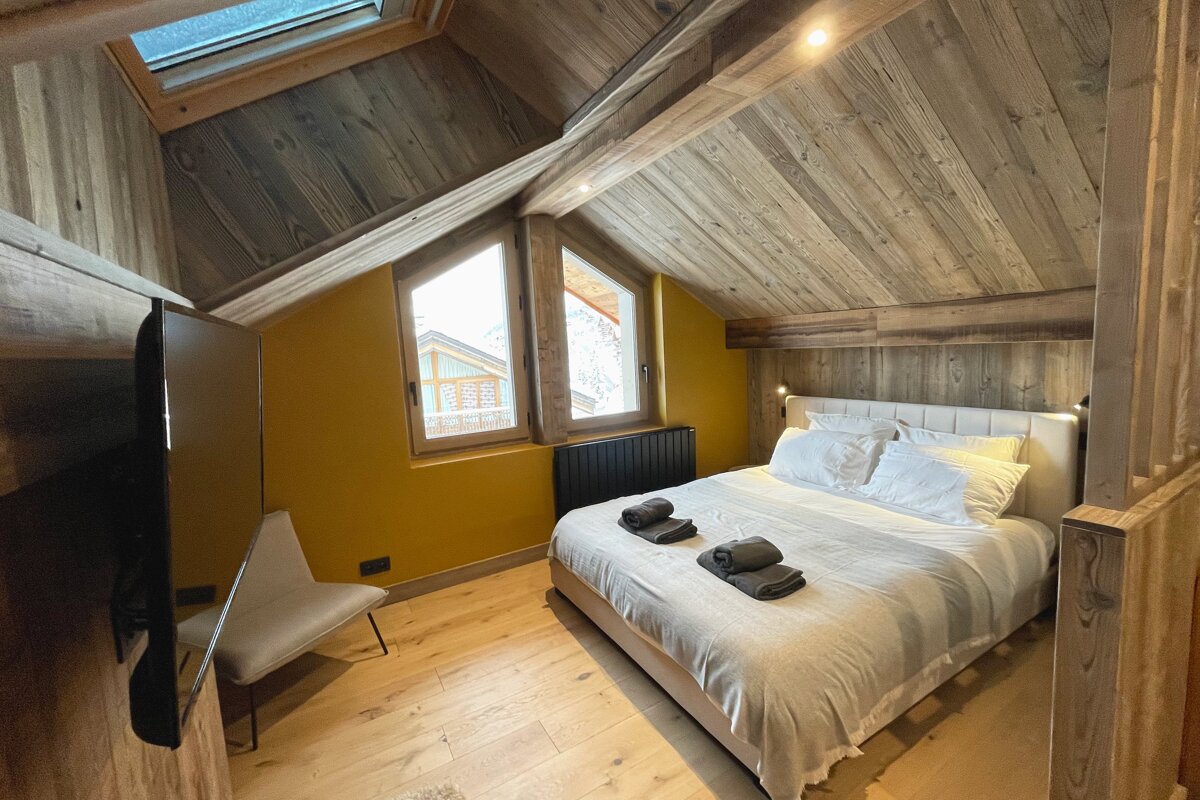 A rustic-modern attic bedroom with wood-paneled walls and ceiling, a large bed with white linens, a yellow accent wall, and bright windows/skylight.