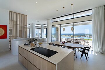 Modern kitchen and dining area with an island, showcasing panoramic views of an infinity pool and lush landscape through expansive windows.