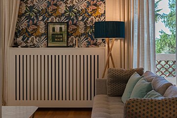 A cozy living room featuring a sofa, a tripod lamp, a white radiator cover, and vibrant tropical wallpaper.
