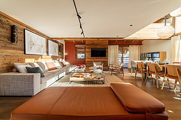 A sunlit, modern rustic living and dining room with warm wood walls, a long sofa, glass coffee table, and a stylish dining set.