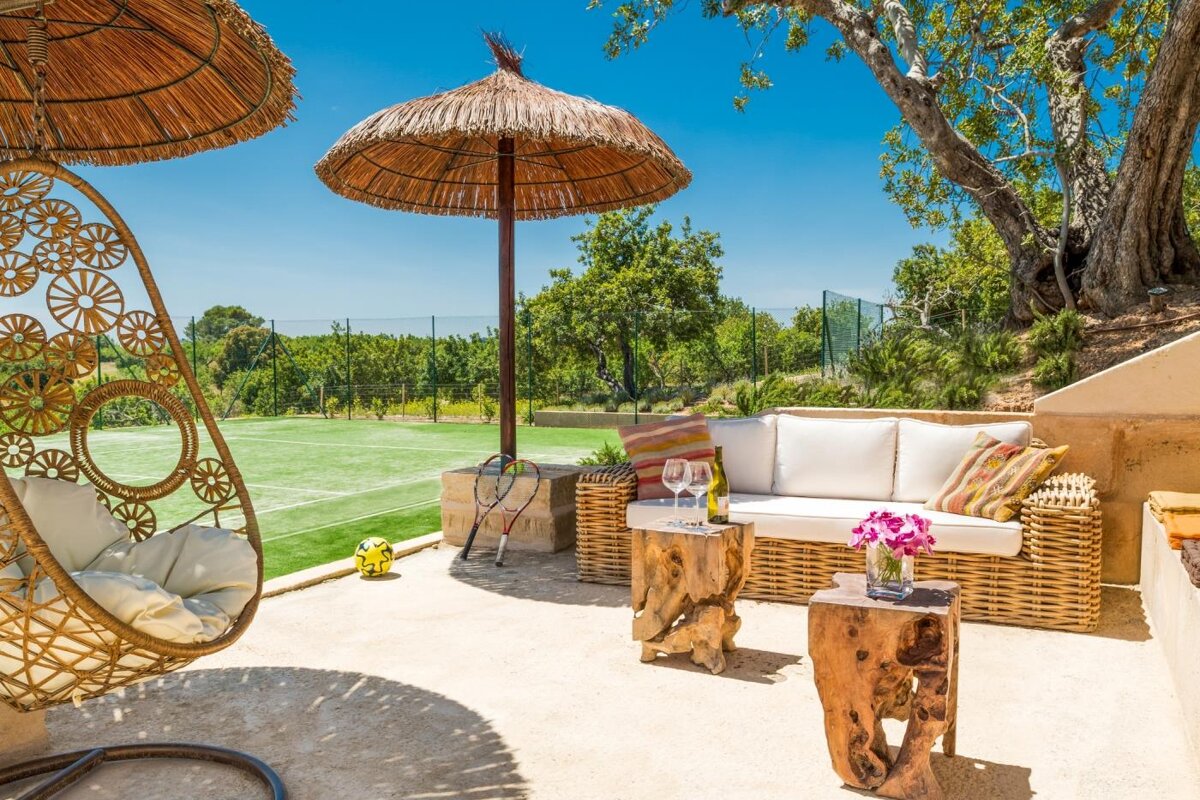 A patio with wicker furniture and umbrellas with a tennis court in the background