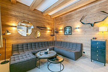 A cozy, modern chalet living room features wood paneling, a grey sectional sofa, nested coffee tables, and warm wall lighting.